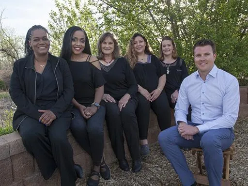 Six dental team members posing outdoors in professional attire against green foliage backdrop.